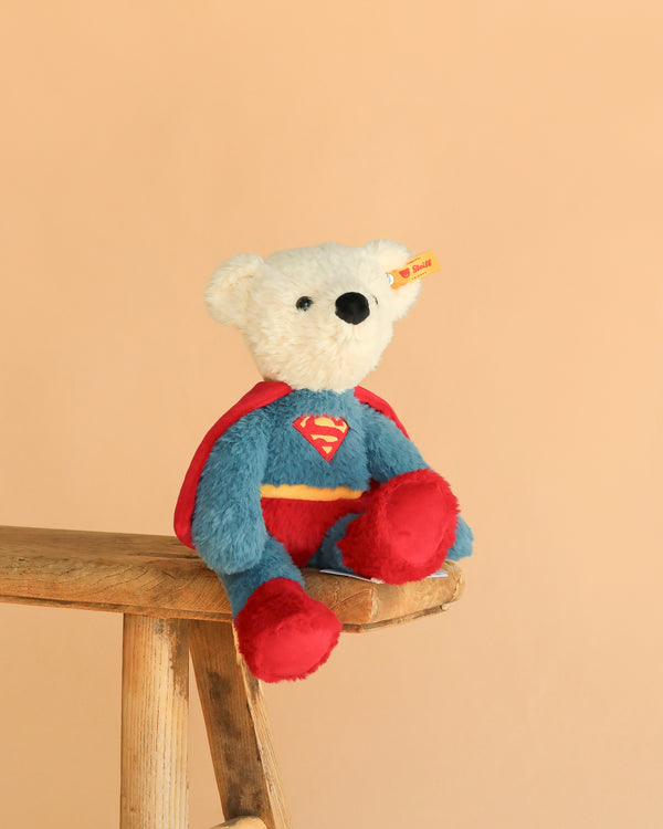The Steiff Superman Teddy Bear, 12 Inches, features a white bear in a blue and red costume with a red cape, sitting on a wooden stool against a beige background—the perfect superhero plush for any fan.