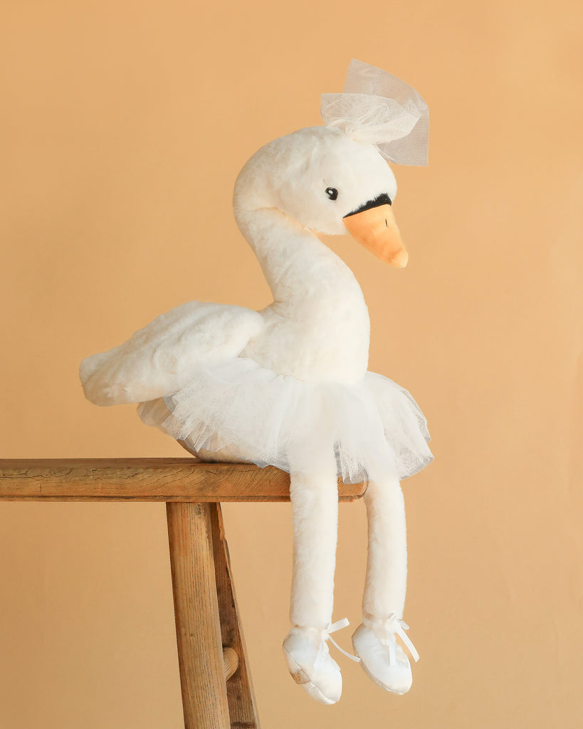 The Bukowski - Odette Ballerina Swan plush wears a white tutu and tulle bow, with long legs and dainty ballet slippers. Posed on a wooden stool against beige, it’s the perfect ballet-themed stuffed animal for any little dancer’s room.