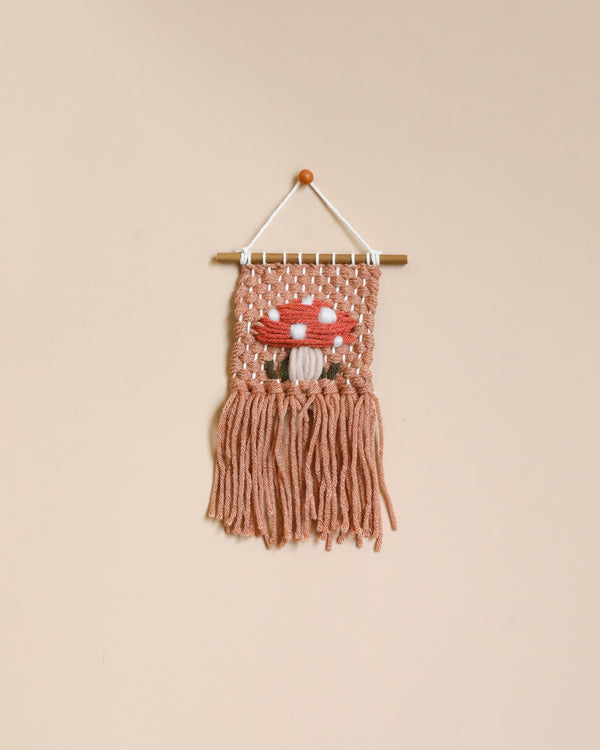 A Handmade Mini Mushroom Wall Hanging - Mauve featuring a mushroom design, crafted from pink and off-white yarns, displayed against a neutral background.