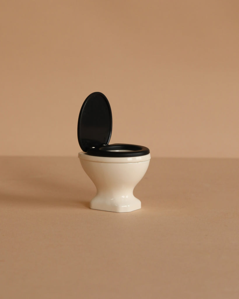 A small, white miniature toilet with a black seat and open lid from the Maileg Suitcasa Furnished Set (ships in one week), displayed on a beige surface with a matching background.