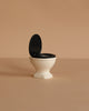 The Maileg Suitcasa Furnished Set (ships in one week) features a small white toilet with a black seat and lid, displayed open on a beige surface—perfect as an accessory for Maileg dollhouses or mouse furniture.