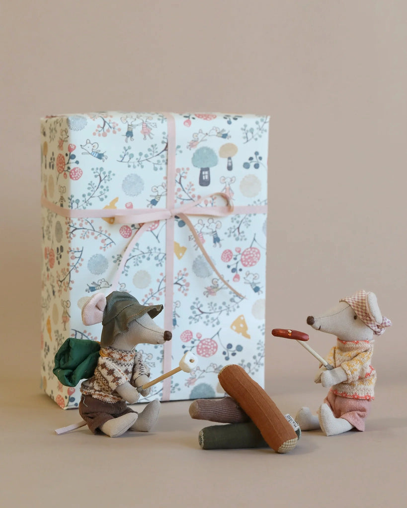 Two small, Maileg Hiker Mice figurines, dressed in colorful camping gear, are sitting beside a Maileg Happy Camper Set - Gift Wrapped, having a conversation. The background is neutral.