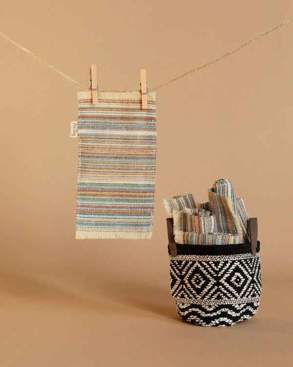 Hanging from a clothesline with wooden clips is a woven multicolored cloth. Below it, a basket with a black-and-white pattern contains several rolled cloths that match the one hanging. Nearby, the Maileg Striped Rug, Medium adds texture to the neutral beige background.