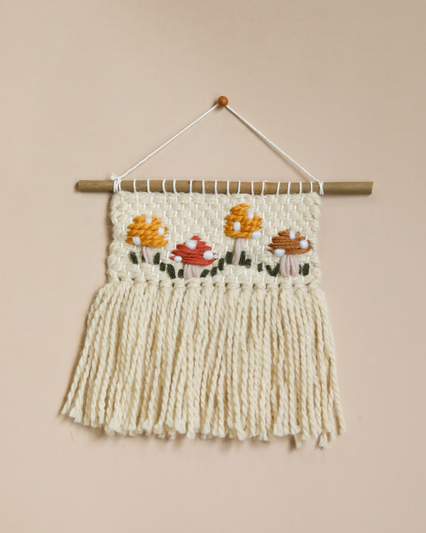 A handmade mushroom wall hanging featuring a floral design with colorful woven flowers on a cream background, ideal as nursery decor, displayed against a pale peach wall.