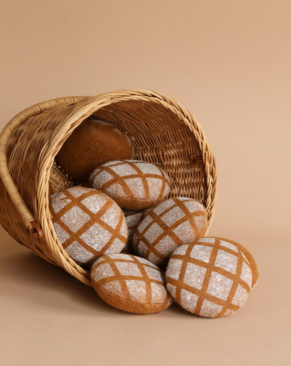 Several freshly baked round handmade Pretend Play Felt Bread loaves with a diamond pattern on top, spilling out of a tipped woven basket against a plain beige background.