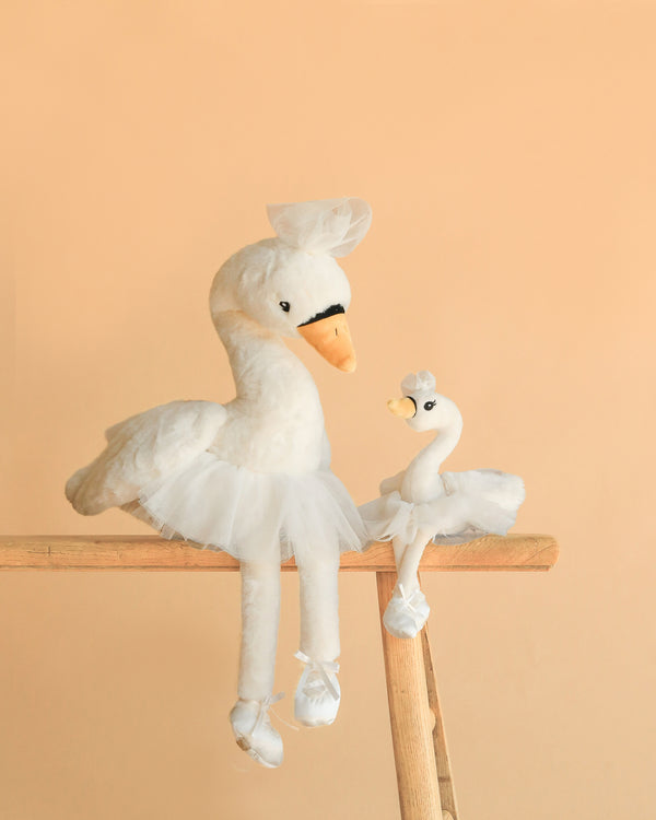 The Bukowski - Odette Ballerina Swan Set features two white plush swans in tutus and tiny hats, one large and one small, posed side by side on a wooden ladder against a peach background, resembling a parent and child.