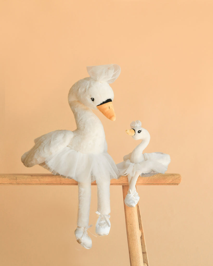 The Bukowski - Odette Ballerina Swan Set features two white plush swans in tutus and tiny hats, one large and one small, posed side by side on a wooden ladder against a peach background, resembling a parent and child.