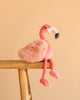 Bukowski - Phoebe Flamingo, a pink plush with a fluffy body, sits on the edge of a wooden stool against a plain beige background—an adorable gift for any plush lover.