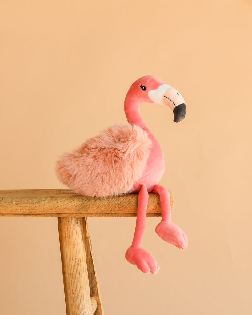 Bukowski - Phoebe Flamingo, a pink plush with a fluffy body, sits on the edge of a wooden stool against a plain beige background—an adorable gift for any plush lover.