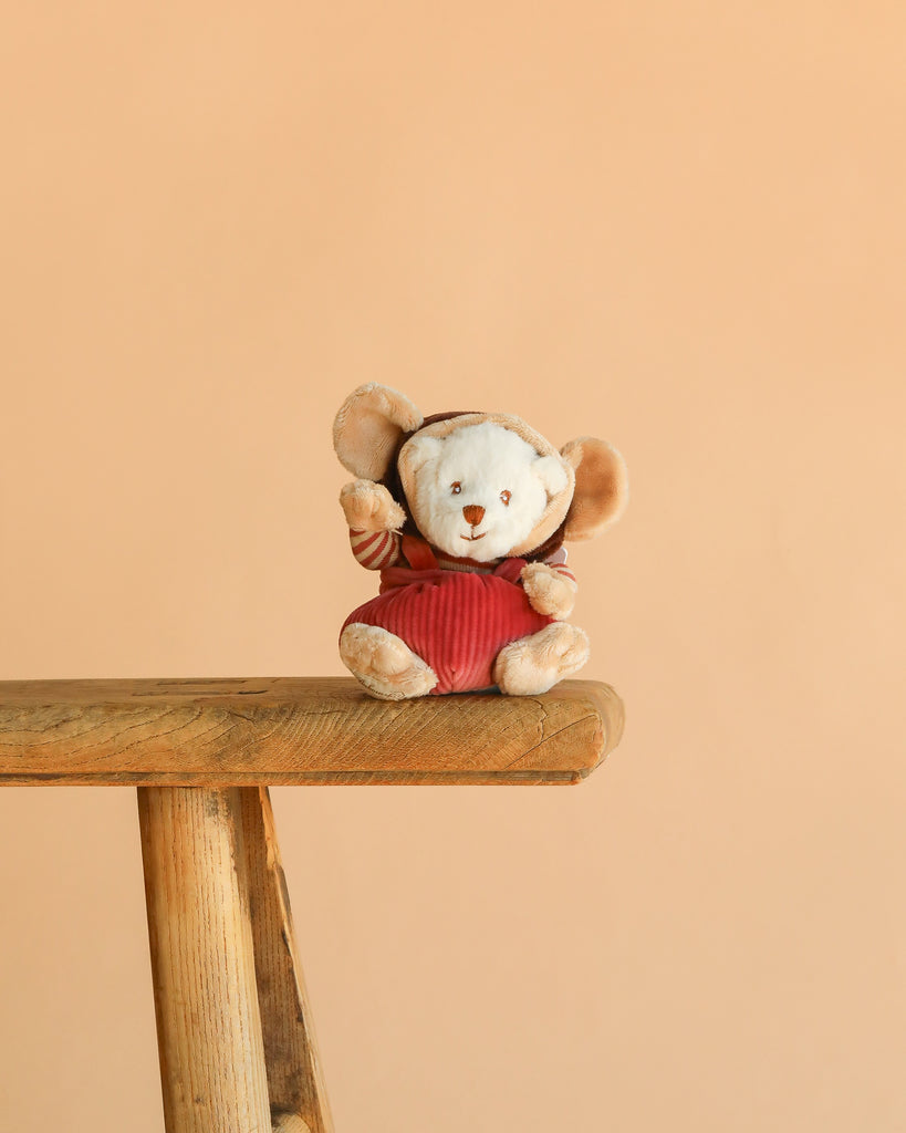A plush Bukowski Ziggy Winston Monkey with large ears and a red outfit sits on a wooden bench against a plain peach-colored background.