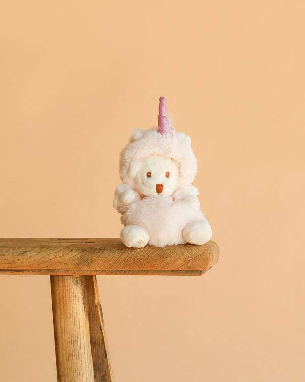 The Bukowski - Ziggy Unicorn is a white plush bear dressed in a fluffy pink unicorn costume with a horn and rainbow mane, sitting on the edge of a wooden bench against a plain peach-colored background.