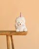 The Bukowski - Ziggy Unicorn is a white plush bear dressed in a fluffy pink unicorn costume with a horn and rainbow mane, sitting on the edge of a wooden bench against a plain peach-colored background.