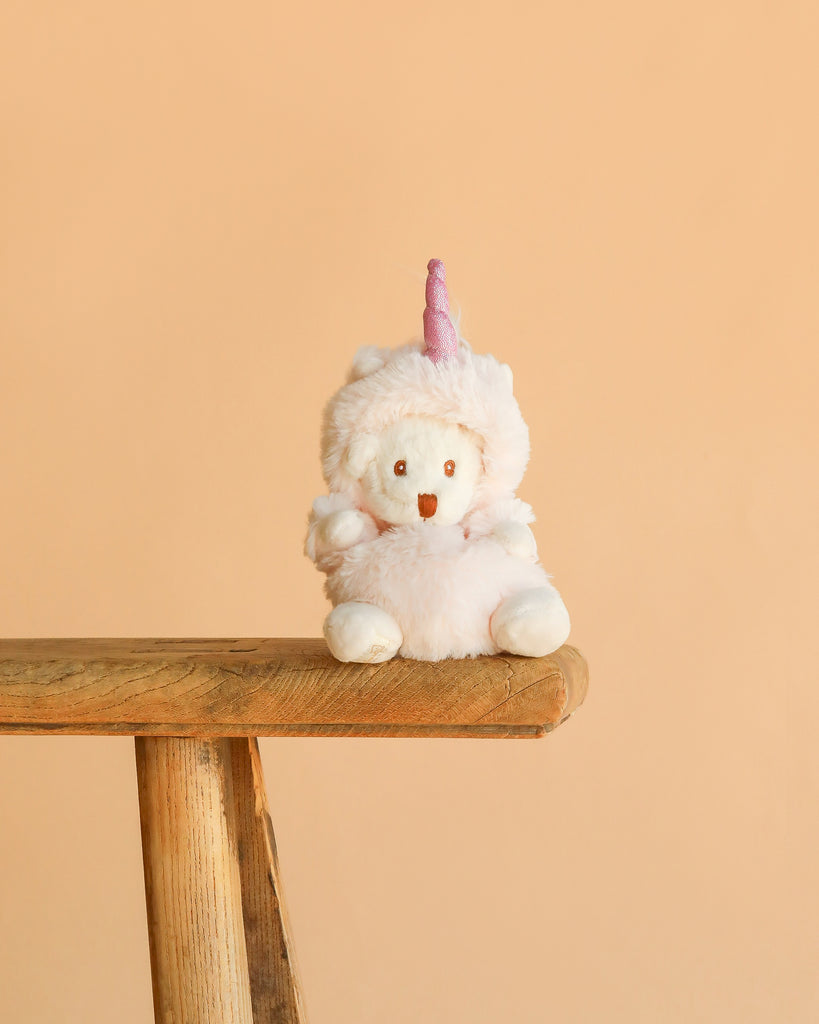 The Bukowski - Ziggy Unicorn is a white plush bear dressed in a fluffy pink unicorn costume with a horn and rainbow mane, sitting on the edge of a wooden bench against a plain peach-colored background.