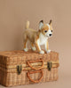 A realistic plush toy model of a Chihuahua dog standing on top of a closed wicker basket with a beige background, crafted from high-quality man-made materials.