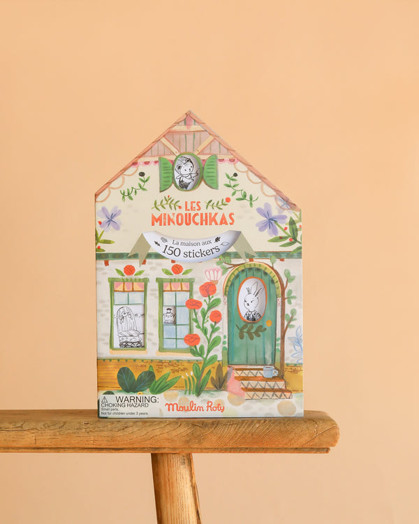 A house-shaped box labeled "Moulin Roty Coloring Book with Stickers - The Minouchkas" features animal and floral designs, contains 150 stickers, and pairs perfectly with any Minouchkas coloring book. Set on a wooden stool against a peach background.