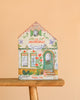 A house-shaped box labeled "Moulin Roty Coloring Book with Stickers - The Minouchkas" features animal and floral designs, contains 150 stickers, and pairs perfectly with any Minouchkas coloring book. Set on a wooden stool against a peach background.