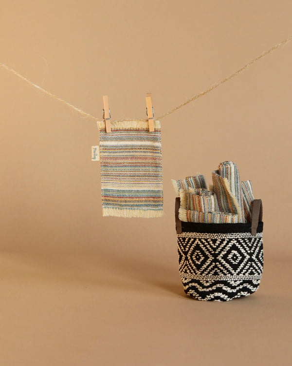 A Maileg Striped Rug, Small is clipped to a clothesline with clothespins. Below it, a black and white patterned basket holds folded textiles that echo the striped designs of the rug, all set against a beige backdrop, creating decor reminiscent of doll houses.