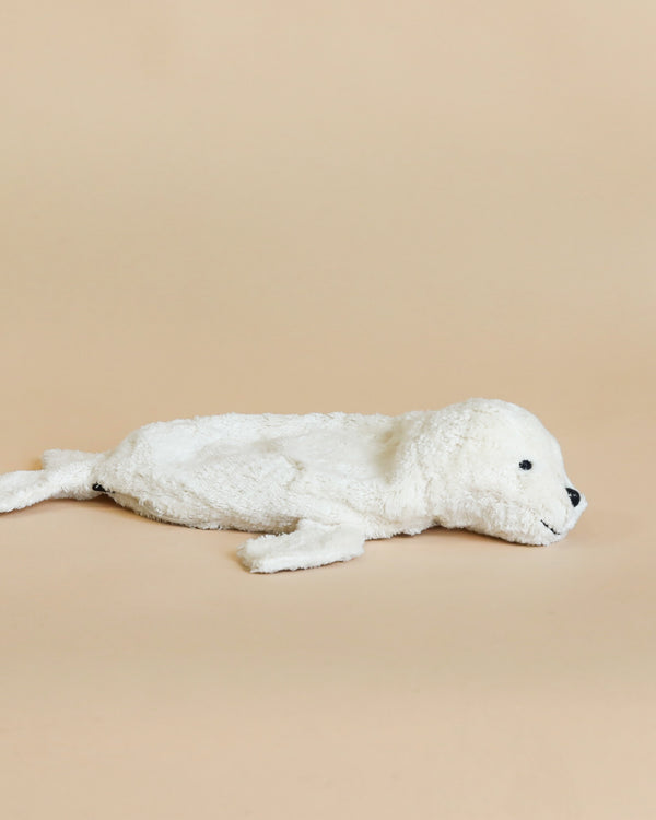 A Senger Naturwelt Cuddly Animal - White Seal lying on a beige background. The seal, handmade in Germany, has a fluffy texture and black eyes, conveying a gentle appearance.