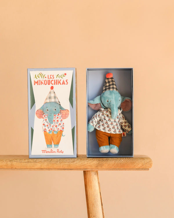The Moulin Roty Pablo the Elephant - The Minouchkas is a boxed toy set with a blue elephant doll in a patterned shirt, brown pants, and plaid hat, shown beside its illustrated box on a wooden surface with a peach background.