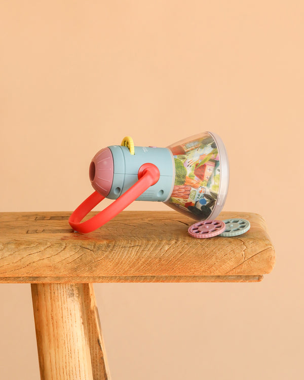 The Moulin Roty Storybook Lantern - The Minouchkas, a vibrant toy projector with a red handle and two round image reels, sits on a wooden surface against a plain beige background.
