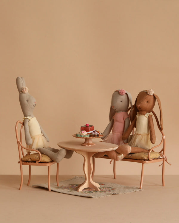 The Maileg Ballet Bunny and Rabbit Set features three dolls in pastel dresses seated around an oval dining table with miniature cakes on vintage-style chairs, set against a beige background for a soft, whimsical effect.