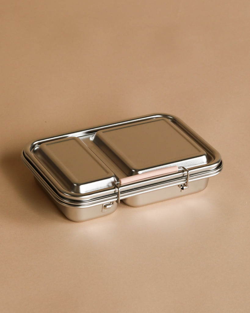 The Haps Nordic Stainless Steel Lunch Box in Blush features two compartments and a secure latch, resting closed on a light brown surface.