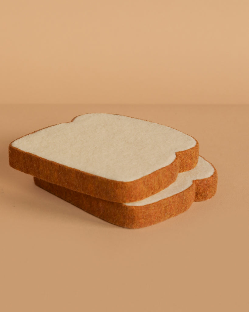 Two slices of felt toast from the Bread and Basket Set are stacked on a beige surface, mimicking real bread with a soft, textured look—ideal for play food or felt breakfast sets.