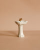 A Maileg Powder Room Set featuring a small, white sink with a gold faucet is centered against a plain beige background.