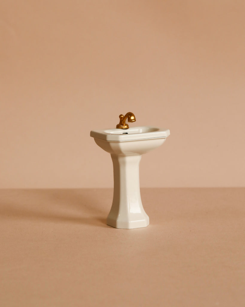 A Maileg Powder Room Set featuring a small, white sink with a gold faucet is centered against a plain beige background.