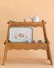 The Moulin Roty Tea Party Suitcase - The Rosalies features a wooden two-tiered stand with a children's tea set—teapot, three teacups with saucers, and a tray decorated with flowers and a rabbit—on a light peach background.