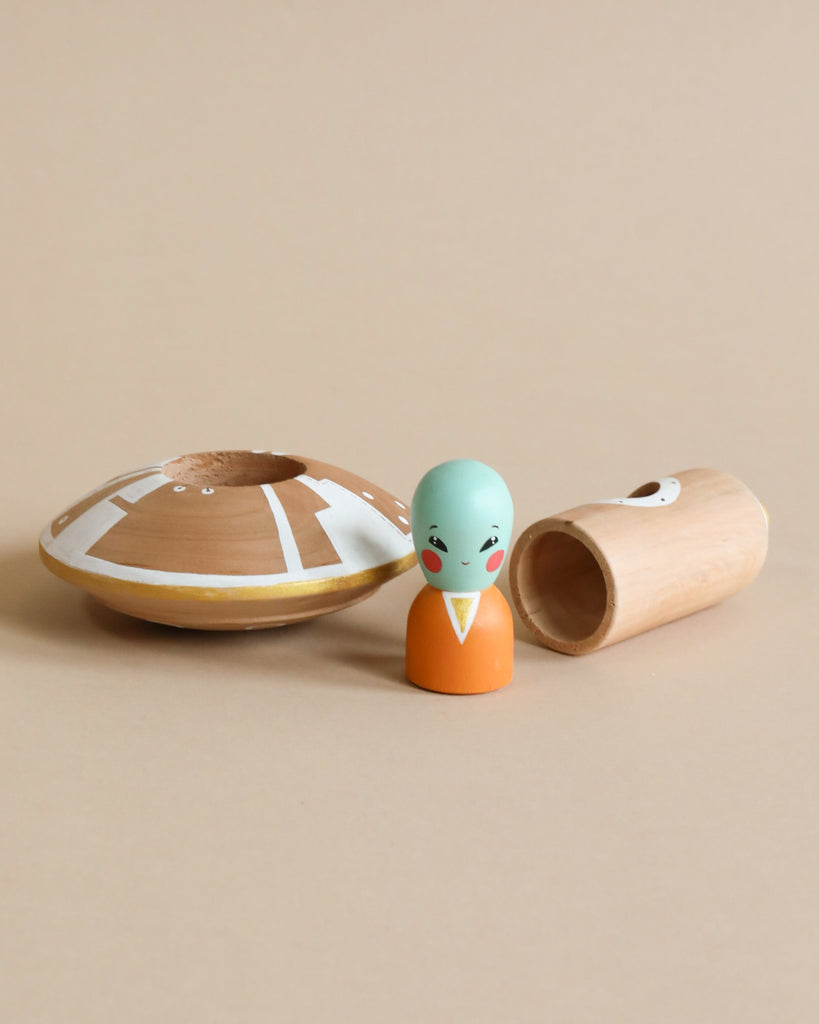 A Handmade Wooden UFO With Alien doll with a mint green face and orange body, standing next to a split bamboo container and a shell-shaped wooden lid on a beige background.