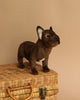 A French Bulldog Dog Stuffed Animal, hand sewn and sitting on a woven wicker basket against a beige background. The toy is brown with detailed facial features and perked-up ears.