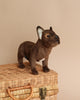 A French Bulldog Beige Dog Stuffed Animal sitting on a woven wicker trunk against a light beige background.
