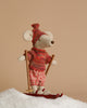 A Maileg Winter Chalet Set features a mouse dressed in a red sweater, hat, and plaid shorts, standing on red skis in faux snow and holding wooden ski poles against a beige background.
