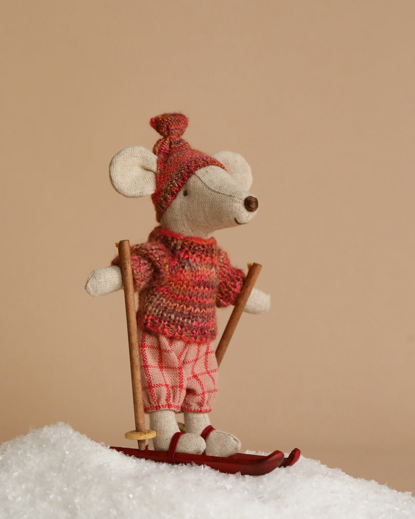 A Maileg Winter Chalet Set features a mouse dressed in a red sweater, hat, and plaid shorts, standing on red skis in faux snow and holding wooden ski poles against a beige background.