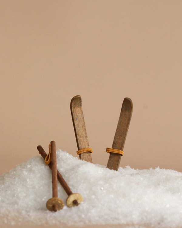 The Maileg Christmas Ski and Ski Poles For Mom & Dad set are partially buried in a small pile of artificial snow against a beige background, creating a playful winter scene.