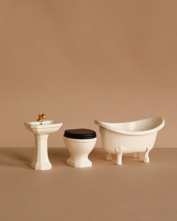 The Maileg Bathroom Set features a pedestal sink with gold faucet, a toilet with black seat, and a clawfoot bathtub, all arranged in a row against a beige background—ideal accessories for your SuitCasa mouse house.