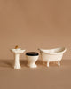 The Maileg Bathroom Set features a pedestal sink with gold faucet, a toilet with black seat, and a clawfoot bathtub, all arranged in a row against a beige background—ideal accessories for your SuitCasa mouse house.