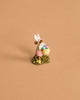 The Wee Forest Folk Tiny Collectible – Hoppy Easter Girl features a handmade mouse figurine in bunny ears and a pink dress, holding a basket of colorful eggs on green grass with yellow flowers.