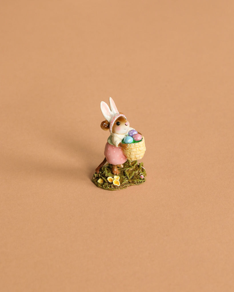 The Wee Forest Folk Tiny Collectible – Hoppy Easter Girl features a handmade mouse figurine in bunny ears and a pink dress, holding a basket of colorful eggs on green grass with yellow flowers.