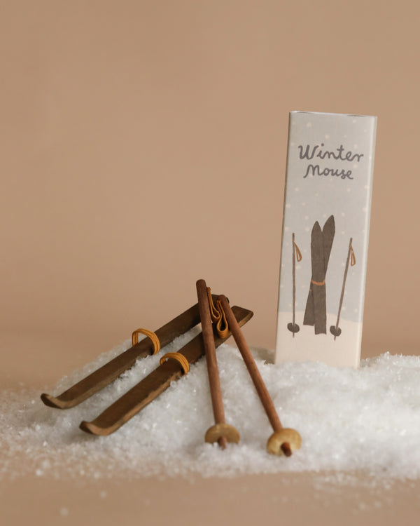 Wooden skis and poles rest on a bed of artificial snow, capturing the essence of mouse winter activity. A rectangular box labeled "Maileg Christmas Ski and Ski Poles For Big Sister & Big Brother," complete with a ski illustration, stands upright beside the display.