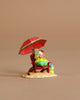 The Wee Forest Folk Tiny Collectible - Perfect Beach Day! shows a figure in green clothes relaxing on a red chair under a red umbrella, reading by a blue bag on sand, all set against a plain beige background.