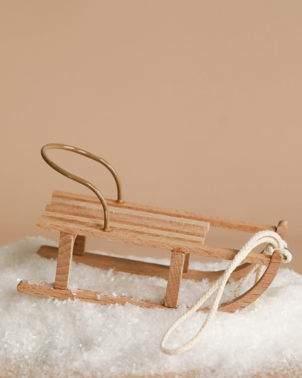 The Maileg Christmas Mini Sled, a perfect winter accessory, rests on a patch of artificial snow against a beige background.