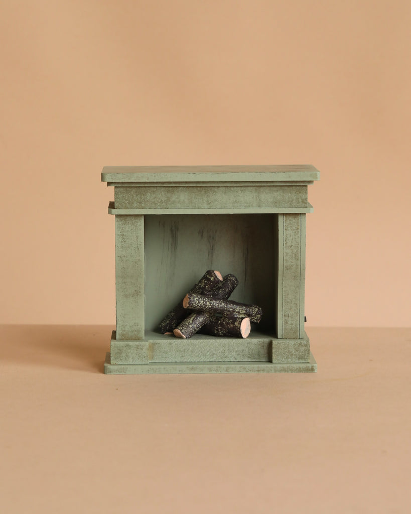 The Maileg Fireplace, Miniature – Dusty Green is a small, green, rectangular wooden fireplace with a vintage finish and stacked logs inside—a charming touch for any miniature home.