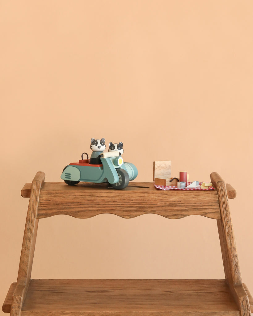 A wooden shelf displays the Badger Picnic Motorbike Ride—a countryside adventure toy motorbike and sidecar with two cat figurines—surrounded by miniature blocks, spools, and fabric on a plain beige background.
