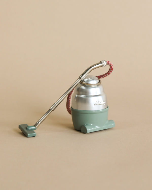 A Maileg Miniature Vacuum Cleaner model in pastel green, displaying "darling" on its front, with a corrugated red hose and a long, thin metal handle, set against a plain