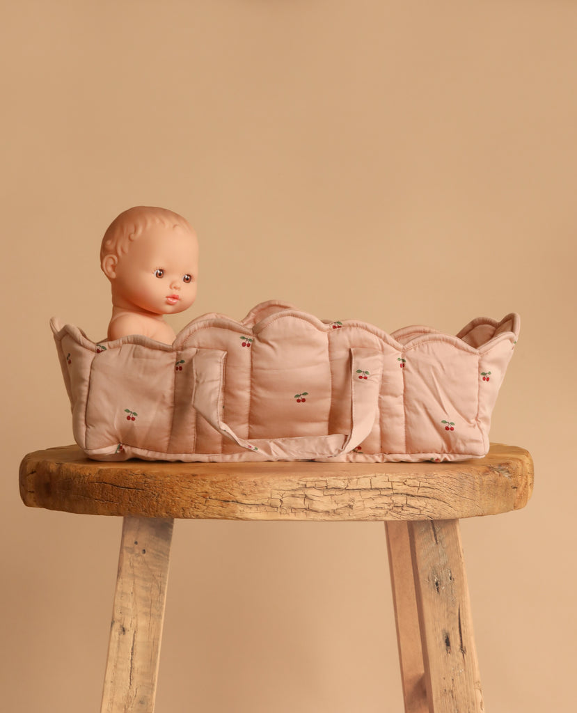A baby doll rests in the Konges Sloejd Doll Lift - Cherry Blush with carry straps, placed on a rustic wooden stool against a beige background.