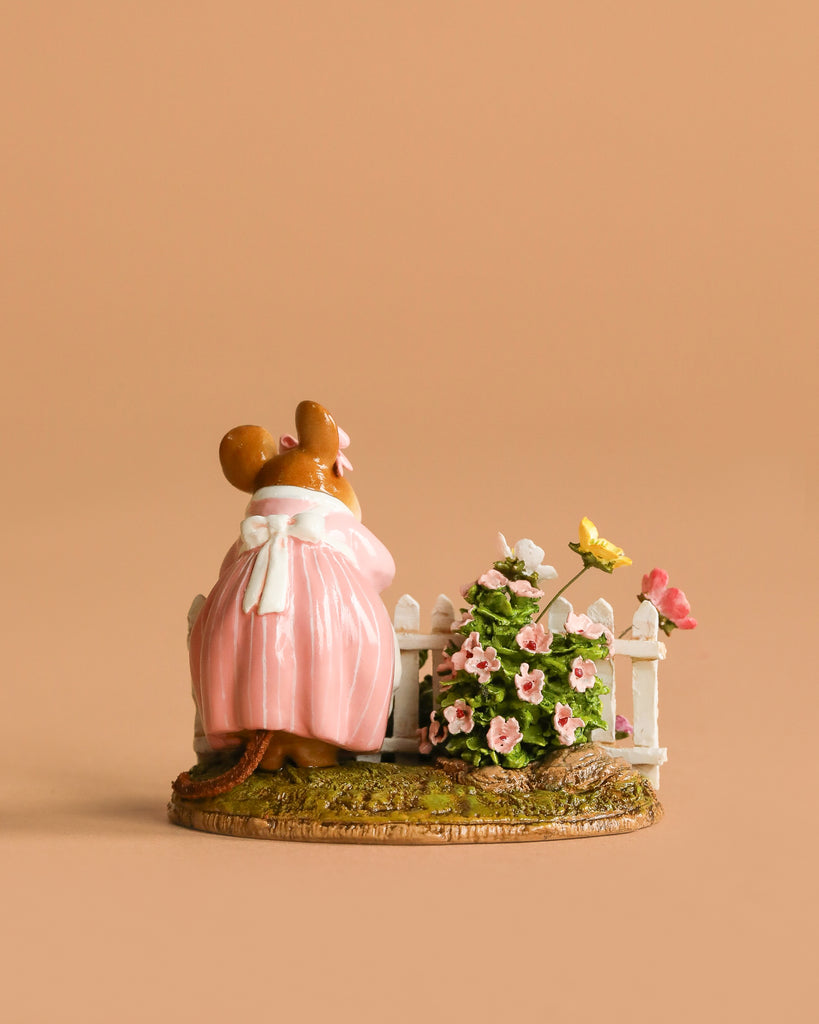 The Wee Forest Folk Tiny Collectible - Annette's Garden features a hand-sculpted mouse in a pink dress beside a white fence with blooming pink flowers and a yellow butterfly, all set against a plain beige background.