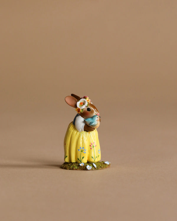 A Wee Forest Folk Tiny Collectible, "Bluebird of Happiness"—a hand-sculpted rabbit in a yellow flowered dress holding a bluebird, with a flower on its head, set on a beige background. Perfect for collectors (Not a Toy).