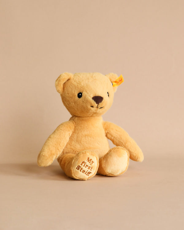 A plush Steiff teddy bear with a soft golden hue sitting against a neutral background. The bear has a tag reading "My First Steiff" on its torso.